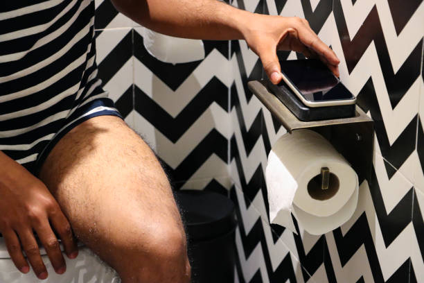 Full frame image of luxury hotel bathroom with unrecognisable man sitting on toilet, putting mobile on phone charging base, toilet roll holder, black and white zig-zag patterned walls and floor tiles Stock photo showing a hotel bathroom featuring a white toilet with modern fittings include bathroom hygiene in the form of a hand shower bidet head attachment, which has been fixed to the wall next to the WC, complete with wall bracket and brass hose connection. pedal bin stock pictures, royalty-free photos & images