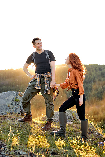 young caucasian couple on hiking trip. enthusiastic man and woman hikers holding hands while walking on mountain trail, in casual clothes, help each other, enjoy travelling together. side view