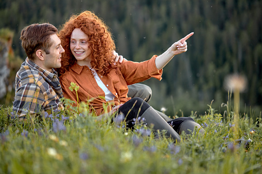 Smiling couple hiking in summer mountains in countryside. Happy woman and man sit on grass. Female and male tourists in casual wear exploring nature, have talk. Side view portrait. Nature, people