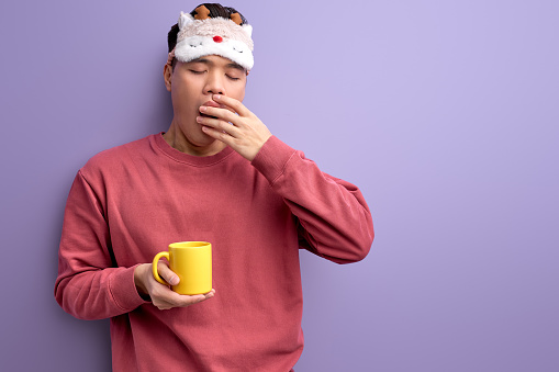 tired asian student with mug in hands yawning, want to sleep before school or university, wearing eyemask for sleep on forehead. isolated purple background