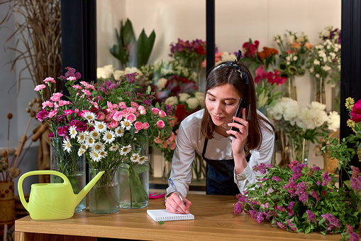 Female attractive florist accepting orders over phone and taking notes in notebook in flower shop. Woman assistant or owner in floral design studio, making decorations. Flowers delivery service.
