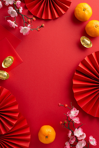 Tradition-rich Chinese New Year display. Vertical top view fans, feng shui decor, coins, sycee, Hong Bao envelopes, tangerines, and sakura arranged on a red surface with frame for text