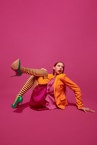 Extraordinary fashion outfit. Young girl dressed in stylish colorful striped orange tights, skirt posing against magenta color background background. Concept of surrealism, art, high, beauty, ad.
