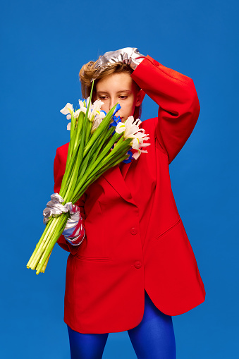 Portrait of weird girl in unusual, strange, variegated colored outfit and posing with flower bunch against color studio background. Concept of high fashion, style and glamour, beauty, love, dating, ad