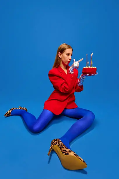 Young lady dressed unusual, freaky, bright outfit, wearing blue tights, red jacket and animal printed heels holding birthday cake and count candles. Concept of high fashion, style and glamour, beauty.