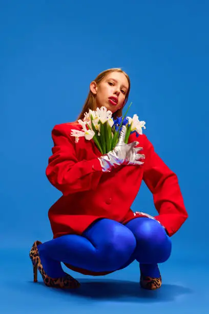 modern fashion. Beauty inside. Weird girl in blue tights with flowers in unusual, strange, red jacket sitting against blue color studio background. Concept of high fashion, style and glamour, beauty.
