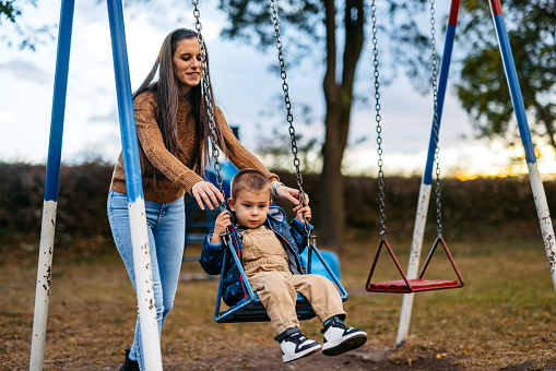 Beautiful young mother pushing her son on the swing in the playground in the park in autumn.