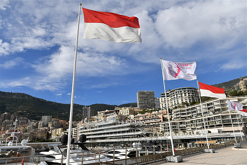Monaco-ville, Monaco: 11 21 2023: Flags of Monaco and the flag to celebrate the centenary of Prince Rainier III fluttering in the wind in front of the luxury yachts in the marina.