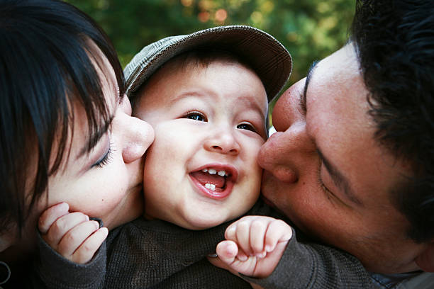 Parents Kissing Child A toddler boy smiles as he gets kisses on his cheeks from his mom and dad. Captured with a Canon 5D Mark II. happy indian young family couple stock pictures, royalty-free photos & images