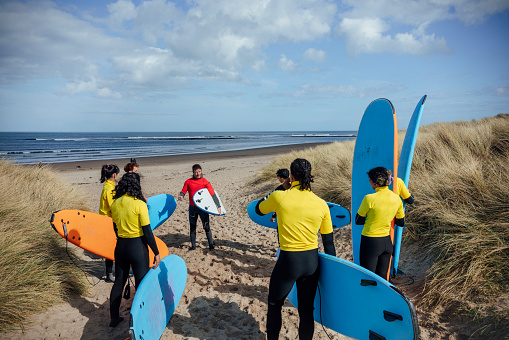 A wide angle rear view of a group of young friends taking part in a surfing lesson on a Beach in Amble in the North East of England. They are stood in front of the instructor who is going over some water and sea safety before they go in.