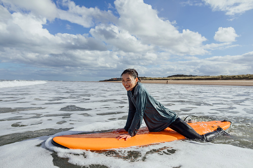 A close-up of a young woman who is taking part in a group surfing lesson in Ambleside in the north-east of England on the coast. She looks delighted to be taking on a new challenge and trying a new hobby. She's riding on a bright orange surfboard and is wearing a wetsuit to keep her warm in the cold north sea.