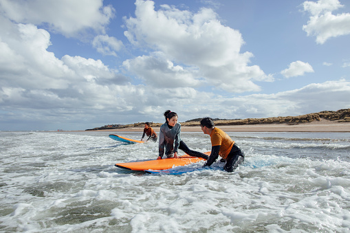 A wide angle side view of a young woman chatting to a new friend, as they take part in a group surfing lesson in Ambleside in the north-east of England, the man she's chatting to is holding his board next to hers and she studies herself on as they enter the water. They are wearing wet suits to keep them warm in the chilly North Sea.