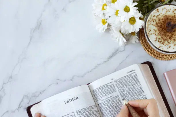 Open holy bible book of Esther with cup of coffee and flowers on white marble background. Top view, copy space. Studying and reading Christian old testament Scriptures.