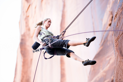 Young Woman Rock Climber at Wall Street near Moab Utah.  Athletic caucasian climber is rappelling down a cliff wearing crop pants and a lime green tank top, she has blond hair with a pony tail.  There is ample copy space in the image for titles and other design elements.