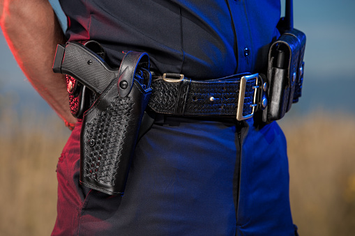 Closeup photograph of the utility belt of a law enforcement officer. A modern handgun in a holster is prominent and in focus in the foreground. Handcuffs, extra magazine clips, and a radio are also visible on the belt.