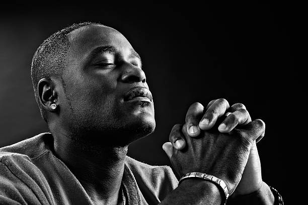 Devout African-American man praying fervently in black-and-white portrait "Black and white portrait of a young African-American man, eyes closed and hands clasped, praying devoutly." church black and white people praying stock pictures, royalty-free photos & images