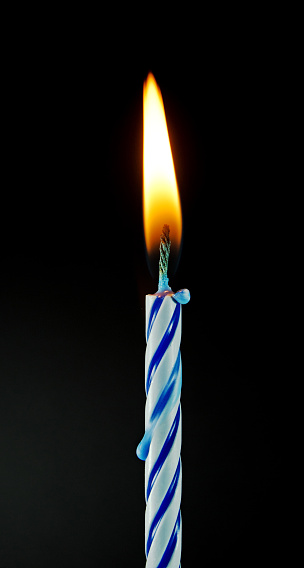 A blue and white birthday candle with a flame on black.