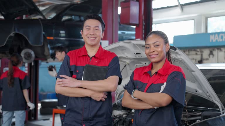 Senior male mechanic and female coworker standing in front of the repairing land vehicle and smiling to a camera with crossarm while two technicians working background. Multiracial people working about motor maintenance at a garage.