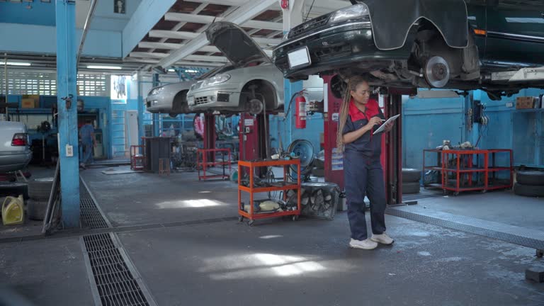 Professional female maintenance engineer inspecting the broken-down vehicle while holding a digital tablet before planning for the repairing procedure in the auto service shop. Young adult engineer working in a garage.