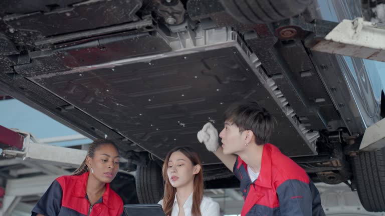 Adult Asian female car repair shop manager explaining the problems with detail and solution on the car to the mechanics while holding a digital tablet. Professional engineers and team working in a garage and repairing a car using hydraulic lift.