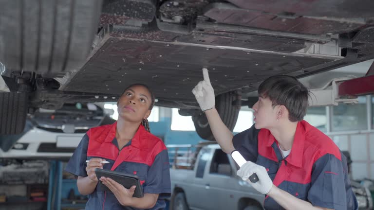 Asian male and African female maintenance engineers examining the undercarriage part of the vehicle lifted up in the garage. Multiracial professional engineers working in auto repair car service for the maintenance and repairing the vehicles.