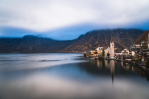 idyllic postcard view of little mountain village of Hallstatt in the austrian mountains, with calm lake, clouds hanging deep over the mountains with last autumn leaf colored trees