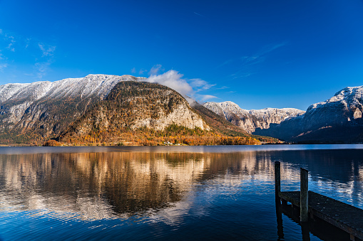 autumn meets winter season on sunny day with blue sky at lake Hallstatt, reflections in the water, small wooden jetty,  colorful larcht rees and some snow in the mountains