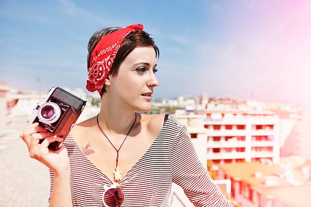 Summer fashion shot of a young woman on the roof of a tall building in the south of France posing with an old-fashioned medium format camera with bellows. Its a beautiful sunny day and the image has visible light leak effects.
