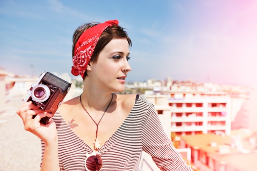 Summer fashion shot of a young woman on the roof of a tall building in the south of France posing with an old-fashioned medium format camera with bellows. Its a beautiful sunny day and the image has visible light leak effects.