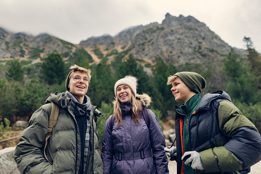 Teenage friends are hiking in the High Tatra Mountains, Slovakia. They are standing by the Popradské pleso lake, talking and laughing. Cold and overcast autumn day.\nShot with Canon R5