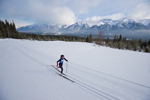 A female cross-country skier goes uphill during a race at the Canmore Nordic Centre Provincial Park in Alberta, Canada. She is doing the classic ski technique. She is wearing a race suit, sunglasses, and a headband.