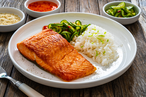 Fried salmon steak with white rice and sliced cucumber on wooden table