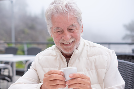 Portrait of attractive smiling bearded senior man sitting outdoors in a foggy day holding an espresso coffee cup.