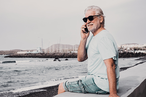 Portrait of relaxed smiling senior bearded man sitting on sea beach at sunrise using mobile phone enjoying freedom and retirement. Elderly male talking on smartphone