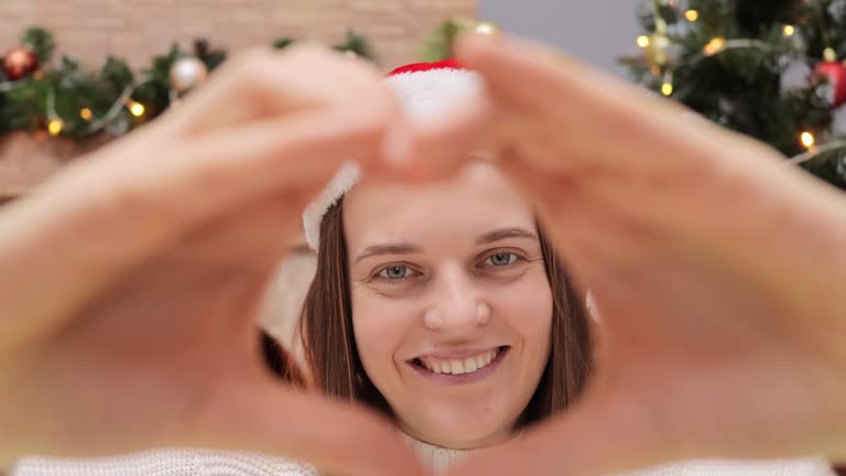 Positive joyful satisfied woman wearing Santa Claus hat looking through heart shape making with hands expressing happiness and love enjoying Christmas atmosphere in decorated room.