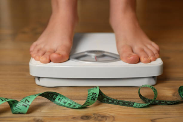 overweight girl using scales near measuring tape on wooden floor, closeup - child obesity imagens e fotografias de stock