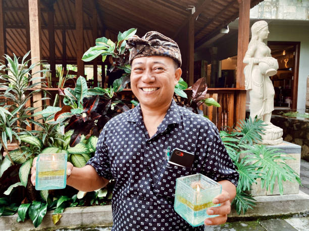 Horizontal closeup photo of a smiling male waiter holding candles in each hand in the garden of a restaurant. Ubud Bali, Indonesia. 2nd January 2020.