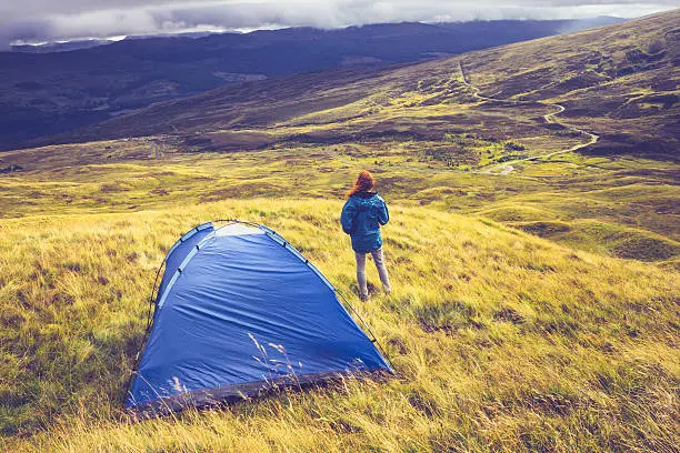 Woman is wild camping and standing next to her tent on a grassy mountain top