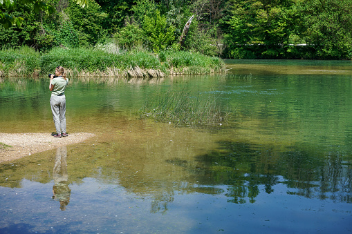 Rear view of one senior woman photographing on the shore of the river against green background.