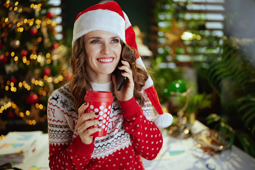 Christmas time. happy elegant small business owner woman in santa hat and red Christmas sweater with cup of hot chocolate speaking on a smartphone in modern green office with Christmas tree.