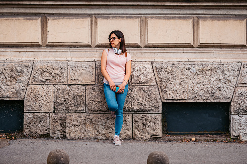 Young woman waiting for a bus while leaning on a building wall.