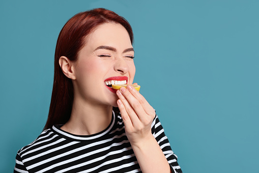 Woman with red dyed hair eating lemon on light blue background, space for text