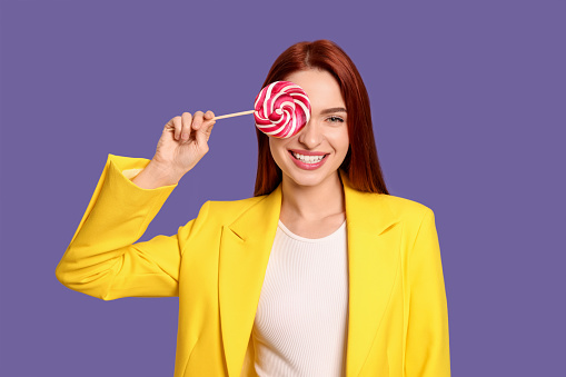 Stylish redhead woman covering eye with lollipop on purple background