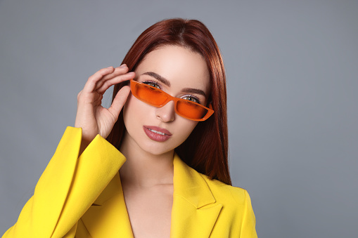 Stylish woman with red dyed hair and orange sunglasses on light gray background