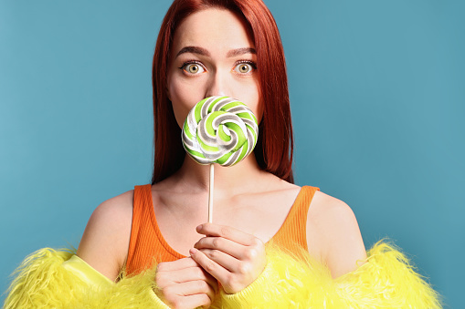 Redhead woman covering mouth with green lollipop on light blue background