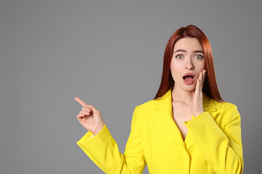 Confused woman with red dyed hair pointing somewhere on light gray background, space for text