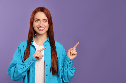 Happy woman with red dyed hair pointing somewhere on purple background, space for text