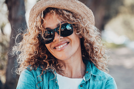 Portrait of young adult woman smiling at the camera with sunglasses and straw hat. Female people happy in outdoors leisure activity. Defocused background with road. Copy space on the right
