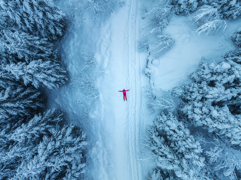 Aerial view of woman lying on the snow on mountain road in beautiful foggy forest in snow in winter day. Top drone view of girl in snowy woods in mist. Pine trees in hoar. Snowfall in wintry woodland