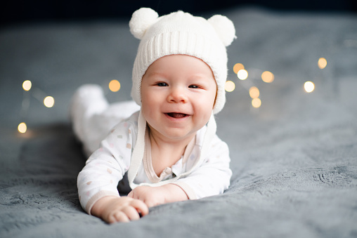 Cute funny infant baby 3-5 months old crawling in bed over xmas lights and wearing knitted hat close up. Winter holiday season. Childhood. Look at camera.